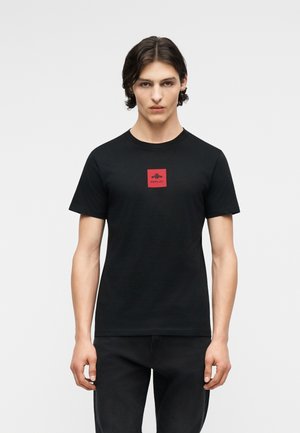 Νέος άνδρας με μεσαίου μήκους σκούρα μαλλιά φοράει μαύρο T-shirt με κόκκινο τετράγωνο λογότυπο και τη λέξη «Replay» στο κέντρο.