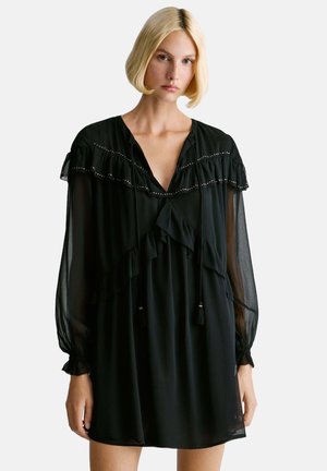 Joven mujer con cabello rubio corto que lleva un mini vestido negro transparente de manga larga con volantes y detalles decorativos.