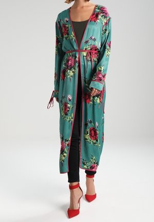 Vrouw draagt een lange turquoise bloemige badjas met roze bies over een zwarte outfit, gecombineerd met rode hoge hakken met enkelriem, staand op een witte achtergrond.