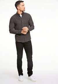 Camicia button-up in tessuto grigio scuro con una texture liscia, dotata di una tasca anteriore, abbinata a jeans neri e sneakers bianche.