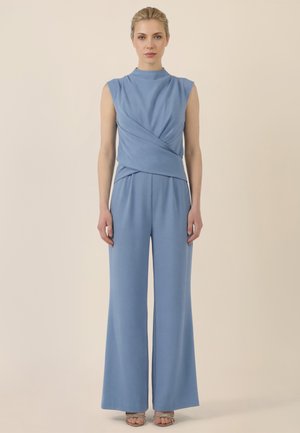 Frau mit blondem Haar, die ein ärmelloses hellblaues Wickeloberteil und passende weit geschnittene Hosen trägt, vor einem beigen Hintergrund stehend.