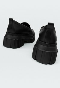 Baskets noires à enfiler avec une semelle épaisse et crantée et un matériau supérieur lisse. Le design présente une couture discrète à l'arrière et aucun matériel visible.