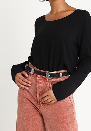 Kvinde iført en sort langærmet skjorte, højttaljede falmede lyserøde jeans og et sort dobbeltspændebælte med sølvdetaljer.