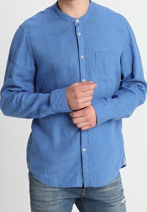 Mężczyzna w niebieskiej koszuli z długim rękawem i stójką, zapinanej na guziki, poprawiający mankiet jednej rękawicy na niebieskich, postarzanych dżinsach.