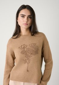 Beige gebreide trui met een ronde hals, voorzien van een textuur motief met bloemenborduursel in een iets donkerdere tint. Ribbelzoom en cuffs.