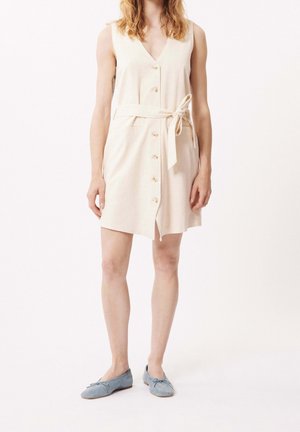 Woman wearing sleeveless beige buttoned dress with tied waist belt and light blue flat shoes against plain white background.