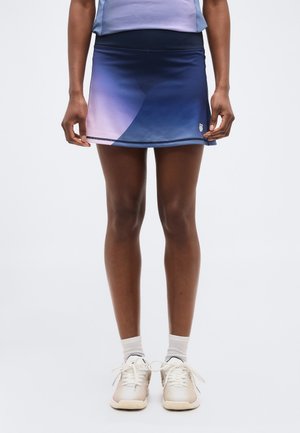 Naine, kes kannab sinise ja lilla üleminekuga spordiseelikut, valgeid pahkluu sokke ja beeže spordijalatsid, seisab tavalise tausta ees.