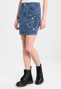 Jeanskjol, blå med blomsterbroderi i rosa och grönt, midja till låren längd, fem fickor, kombinerad med svarta snörstövlar.