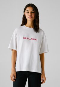 Ung kvinde med mørkt hår iført en hvid oversized T-shirt med rød "RED BULL RACING" tekst og sorte bukser, stående foran en ensfarvet baggrund.