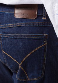 Vaqueros de denim en azul oscuro con costuras doradas. Presenta una etiqueta de cuero en la parte posterior y formas de bolsillo distintivas con costuras en ángulo.