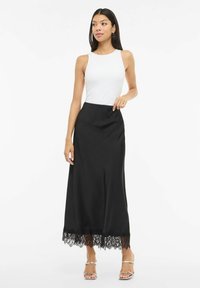 Vrouw die een witte mouwloze top draagt, een zwarte midi-rok met kanten zoom en zilveren sandalen met bandjes, staand tegen een witte achtergrond.