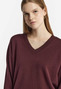 Burgunderfarbener, locker sitzender Pullover mit V-Ausschnitt, aus weichem Material gefertigt. Mit überschnittenen Schultern und einem strukturierten Strickdesign.