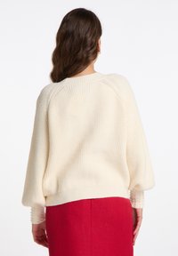 Pulover tricotat de culoare crem deschis, cu textură cu nervuri, umeri lăsați și mâneci transparente, cu aplicații. Purtați-l cu o fustă roșie.