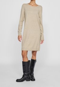 Abito maglione beige con maniche lunghe, polsini e orlo a coste; abbinato a stivali neri in pelle al ginocchio con dettaglio a fibbia.