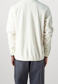 Pullover bianco in fleece con maniche lunghe e scollatura a V, caratterizzato da una vestibilità morbida e una trama sottile; abbinato a jeans grigio scuro.