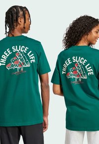 Vihreät puuvillapaita, joissa on iso pizzakuvasto punaisilla täytteillä ja teksti "THREE SLICE LIFE" selässä. Lyhyet hihat, rento malli.