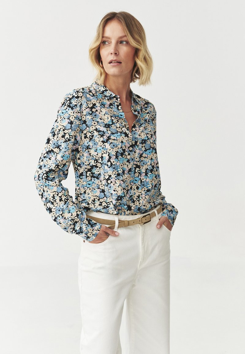 Cămașă cu imprimeu floral, cu butoni, având un model negru, albastru și crem, cu mâneci lungi și guler, asociată cu pantaloni albi, cu talie înaltă.