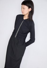 Schwarzes Kleid mit einem Reißverschluss vorne, langen Ärmeln und einer gerafften Taille. Der Stoff hat eine glatte Textur mit horizontalen Rippdetails.
