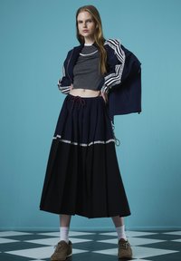 Marineblauwe jas met gestreepte mouwen, grijze crop top en zwarte plissérok met kanten accenten; bruine schoenen; tegen een blauwe achtergrond.