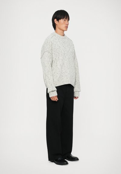 Langermet, lys grå strikkegenser med et teksturert mønster, kombinert med løstsittende svarte bukser og svarte slip-on sko.
