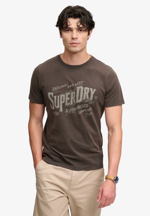 Junger Mann mit dunklem lockigem Haar, der ein braunes Superdry-T-Shirt, beige Hose und eine braune Lederarmbanduhr an seinem linken Handgelenk trägt.