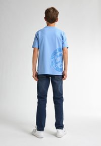 Camiseta de algodón de color azul claro con un logo circular impreso en la parte de atrás. Combinada con jeans azul oscuro y zapatillas blancas.