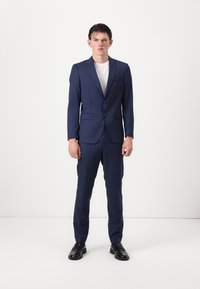Marineblauer Anzug mit einem taillierten Blazer, der über zwei Knöpfe und einen Reverskragen verfügt, kombiniert mit passenden Hosen und schwarzen Schuhen. Darunter ein weißes T-Shirt.