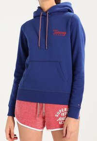 Felpa blu con cappuccio, dotata di tasca a marsupio frontale e logo rosso ricamato. Indossata con pantaloni corti rossi testurizzati con dettagli bianchi.