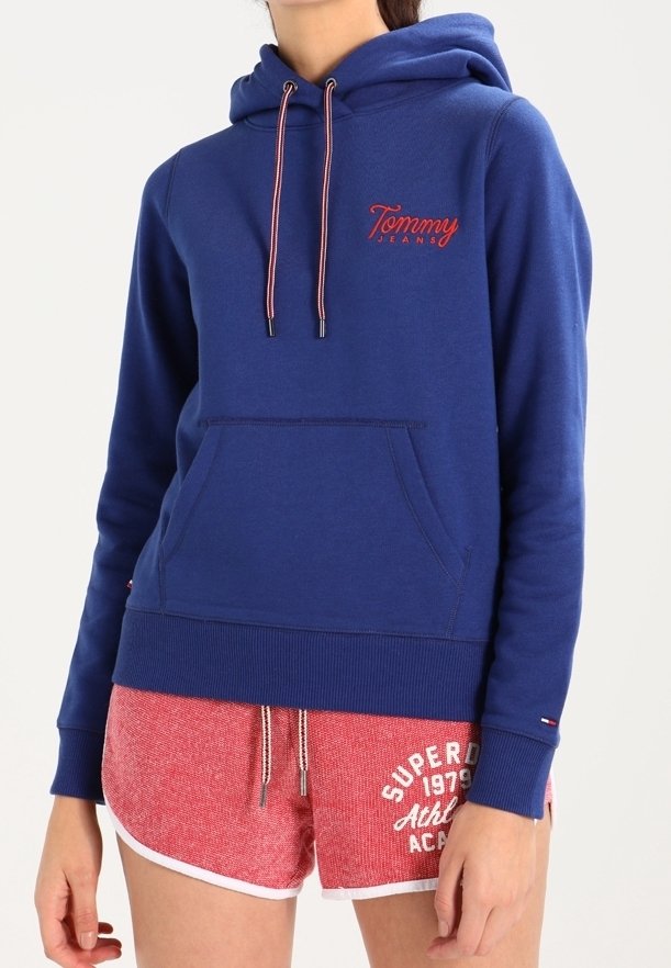 Felpa blu con cappuccio, dotata di tasca a marsupio frontale e logo rosso ricamato. Indossata con pantaloni corti rossi testurizzati con dettagli bianchi.