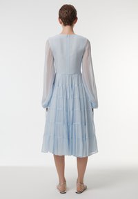 Robe bleu clair en couches avec de longues manches transparentes et un col rond. Présente une fermeture éclair dissimulée au dos et un tissu texturé.