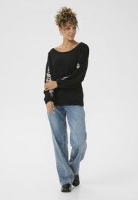 Donna con capelli ricci che indossa un top nero a maniche lunghe con ricami floreali bianchi e jeans blu larghi, in piedi con le braccia incrociate.