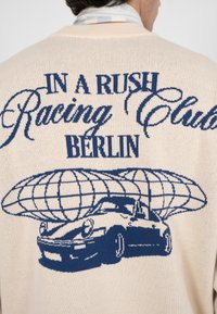 Pull beige en maille avec le texte graphique bleu "IN A RUSH Racing Club BERLIN" et un design stylisé de voiture avec un globe au-dessus.