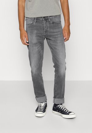 Person trägt hochgekrempelte graue Jeans und schwarze Sneaker mit niedriger Schafthöhe und weißen Schnürsenkeln, steht vor einem schlichten hellen Hintergrund.