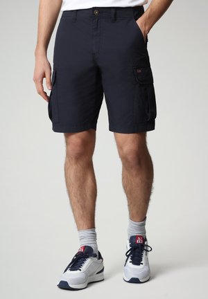 Homme portant un short cargo bleu marine, des chaussettes blanches et des baskets blanches aux lacets bleu marine, debout avec une main dans la poche sur un fond neutre.