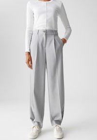 Light gray pleated trousers with a high waist, button closure, and side pockets. Paired with a fitted white long-sleeve top and white sneakers.