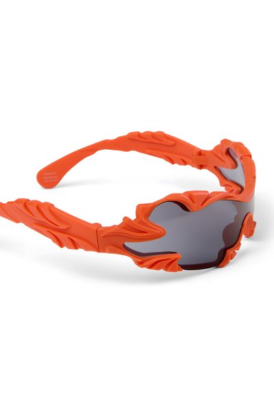 Sportliche orangefarbene Sonnenbrille mit gezackten, skulptierten Rahmenkanten und dunklen, umschließenden Gläsern vor weißem Hintergrund.