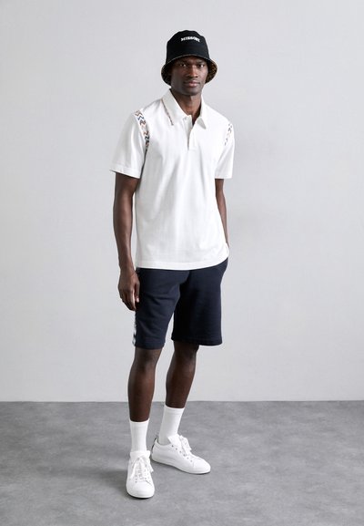 Λευκό πόλο μπλουζάκι με πολύχρωμες γραμμές σε ζιγκ ζάγκ στους ώμους, σε συνδυασμό με ναυτικές βερμούδες, λευκά αθλητικά παπούτσια και ένα μαύρο καπέλο τύπου bucket με λογότυπο.