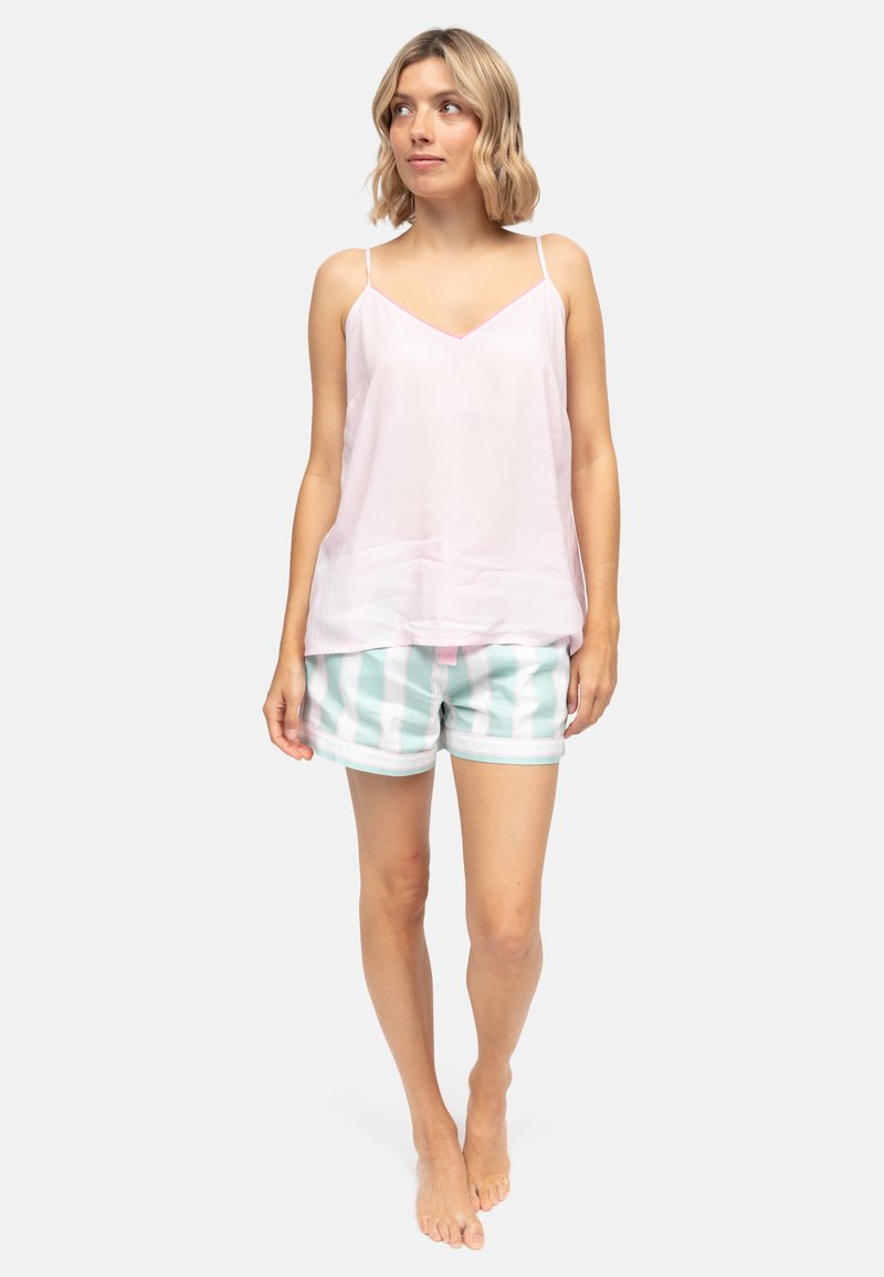 Młoda kobieta stojąca boso, ubrana w jasnoróżową, bezrękawową bluzkę i miętowo-białe krótkie spodenki w paski, patrząca w bok.