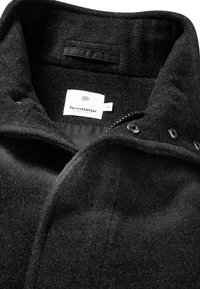 Schwarze Jacke aus Wollmischung mit hohem Kragen, Knopfverschluss und einem inneren Etikett der Marke "hessnatur". Texturierte, weiche Stoffqualität.