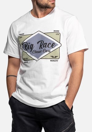 Homme portant un T-shirt blanc avec le graphisme "Big Race Classic Cars" et un short noir, mains dans les poches, légèrement tourné vers la gauche.