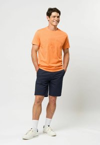 Oranžinė medvilninė marškinėliai su U.S. Polo Assn. logotipu, derinami su tamsiai mėlynomis šortais. Avalynė - baltos sportinės bateliai su mėlynais akcentais ir balti kojinės.