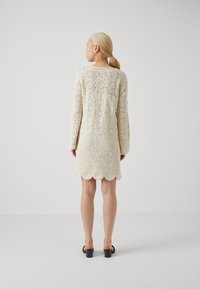 Robe au crochet en crème, avec des motifs circulaires, des manches longues et un ourlet festonné, accompagnée de sandales à talons ouverts noires.
