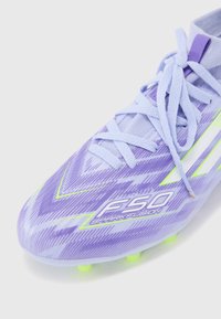 Crampons de football violets avec un motif texturé, présentant des accents verts, un logo sur le côté et des lacets bleu clair. Éléments en caoutchouc sur la semelle.