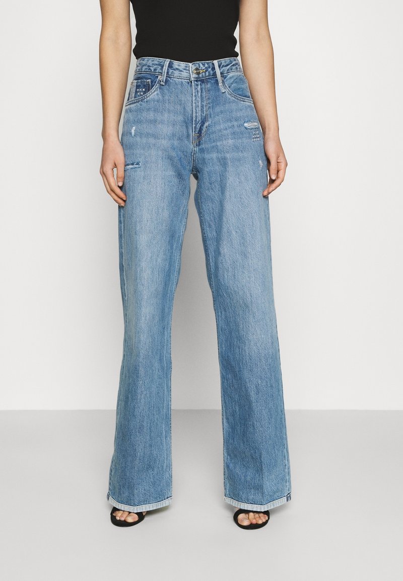 Femme portant un jean denim bleu clair taille haute, coupe large avec une légère usure, associé à un top noir sans manches et des sandales noires à bouts ouverts.