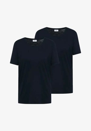 Frau steht vor einer beigen Wand und trägt ein marineblaues T-Shirt, cremefarbene weite Hosen, weiße Sneakers und eine olivgrüne Umhängetasche.