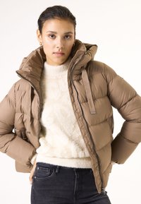 Chaqueta tipo plumón marrón con capucha, que presenta un diseño acolchado, cierre de cremallera y un suave suéter beige debajo. Tejido texturizado.