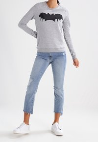 Persona che indossa un felpa grigia con una grafica di un pipistrello nera, jeans cropped strappati di colore blu chiaro e sneakers bianche, in piedi su uno sfondo bianco.