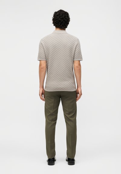 Άνδρας, ορατός από πίσω, φοράει κοντομάνικο πόλο σε ανοιχτό γκρι με υφή, ελαιχρώμο παντελόνι και μαύρα παπούτσια, στέκεται μπροστά σε λευκό φόντο.