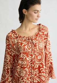 Ruchovaná off-shoulder blúzka v hrdzavej farbe s krémovým paisley vzorom. Obsahuje viazanie na výstrihu a rozšírené rukávy. Hladká textúra látky.