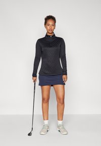 Černý prošívaný top s dlouhým rukávem a zipovým límcem, spárovaný s námořnickou golfovou sukní. Drží golfovou hůl, na nohou má béžové golfové boty.